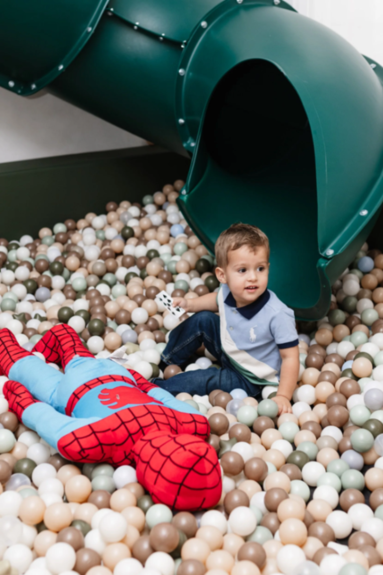 A toddler sitting in the ball pit beneath the green tube slide.