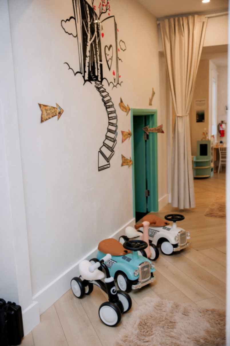 Ride-on cars parked beside the mural wall and play doorway inside the Play Room.